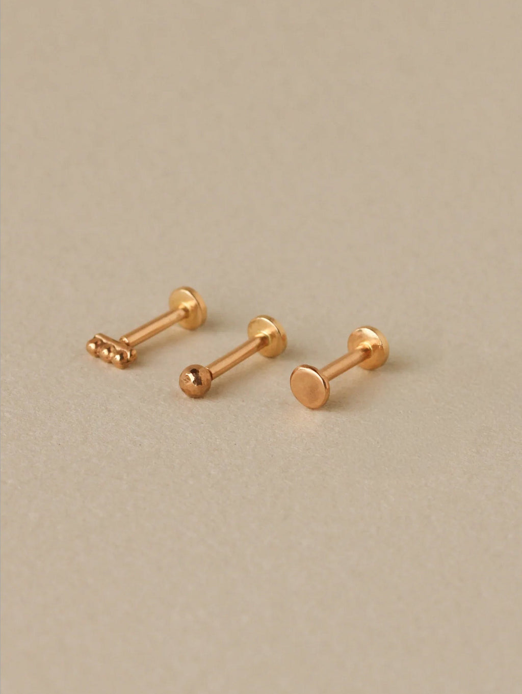 Three gold earrings with a small decorative front and a flat back on a beige background